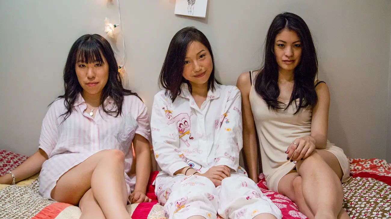 The three leading women of 'Flat 3' sit in their pyjamas on a bed.